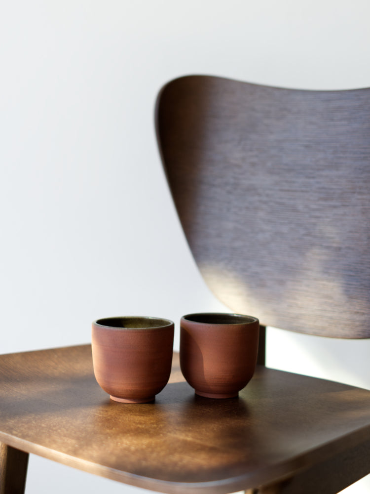 Twee handgemaakte kopjes van rode klei op een bruine houten stoel