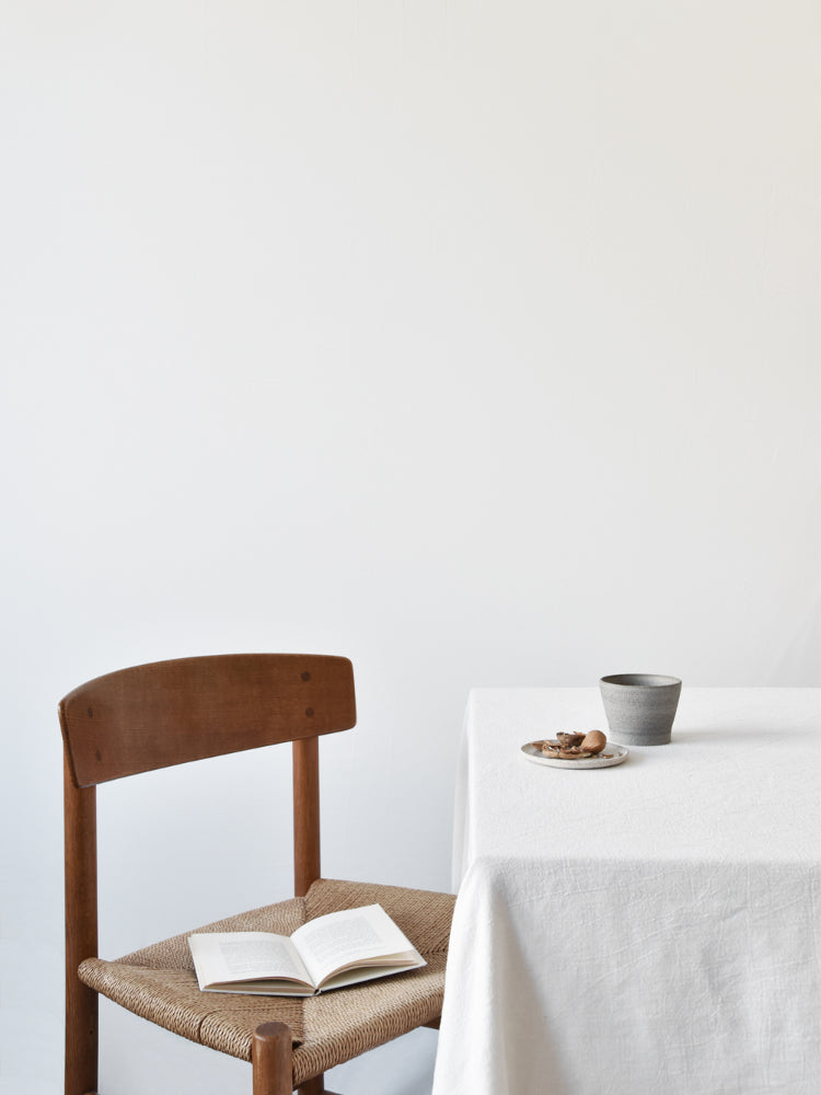 Handgemaakt bruin keramieken kopje op tafel met stoel ernaast en opengeslagen boek slow living