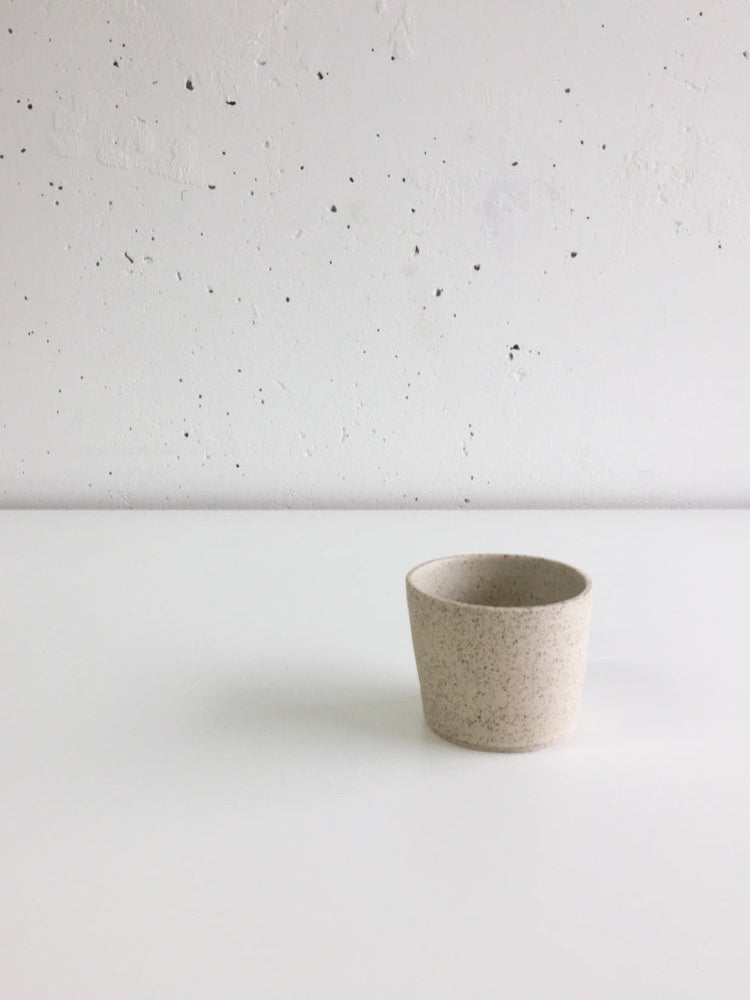 Handgemaakt kopje van lichtgespikkelde klei op een witte tafel met een witte muur als achtergrond