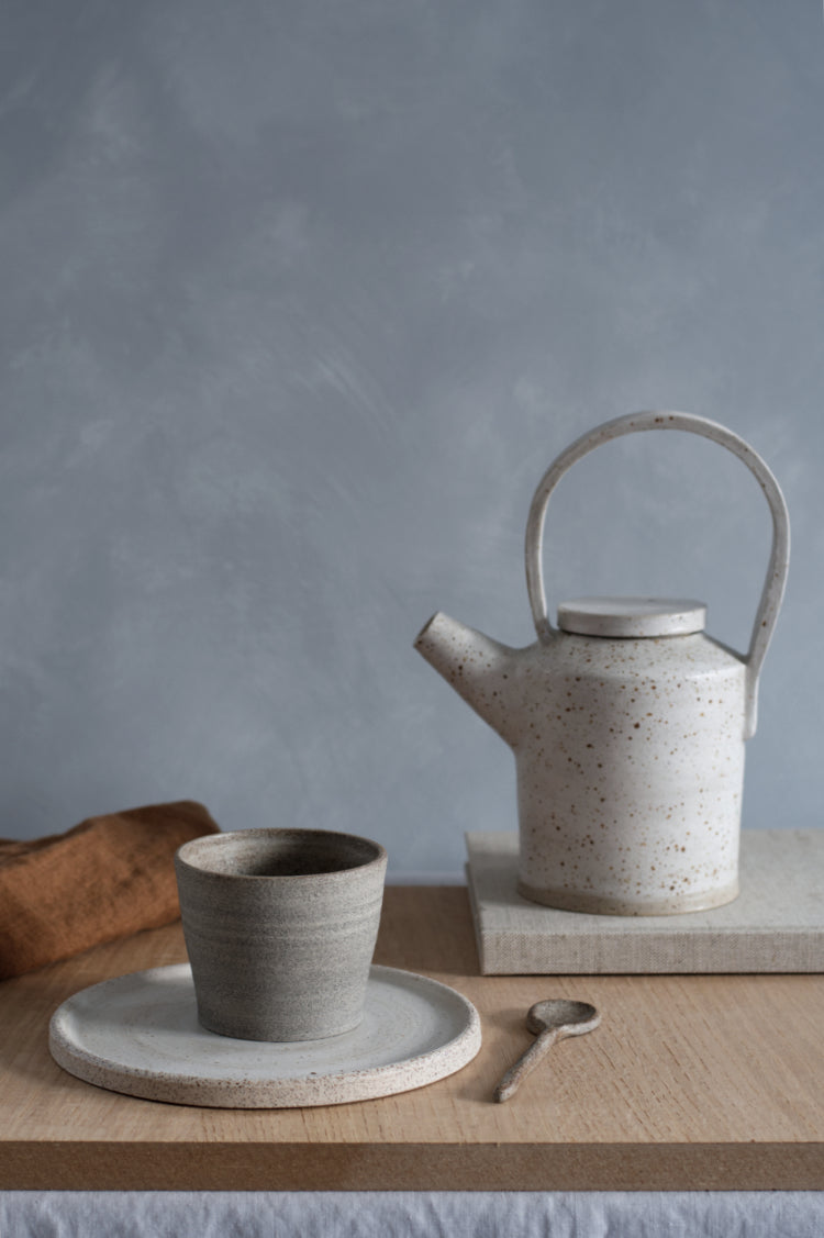 Rustige tafelscène: een donker kopje en theepot op een wit gespikkeld keramisch bord.