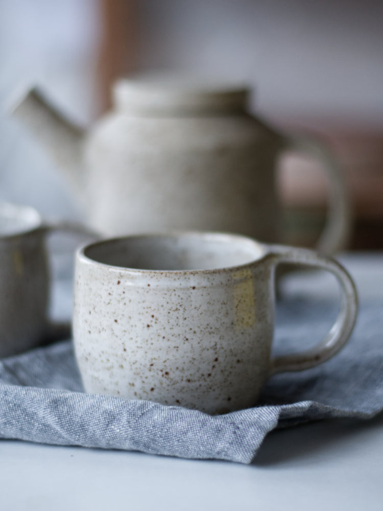 Handgemaakte mok wit gespikkeld glazuur op een blauwe theedoek, een theepot vaag in de achtergrond