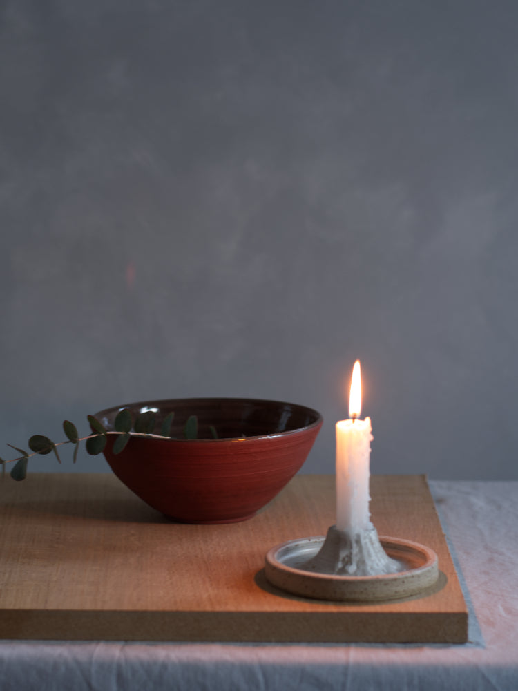 Handgemaakte kaarsenhouder van gespikkeld keramiek met een brandende kaars erin, erachter een kom van rode klei met een takje erin, slow living vibe