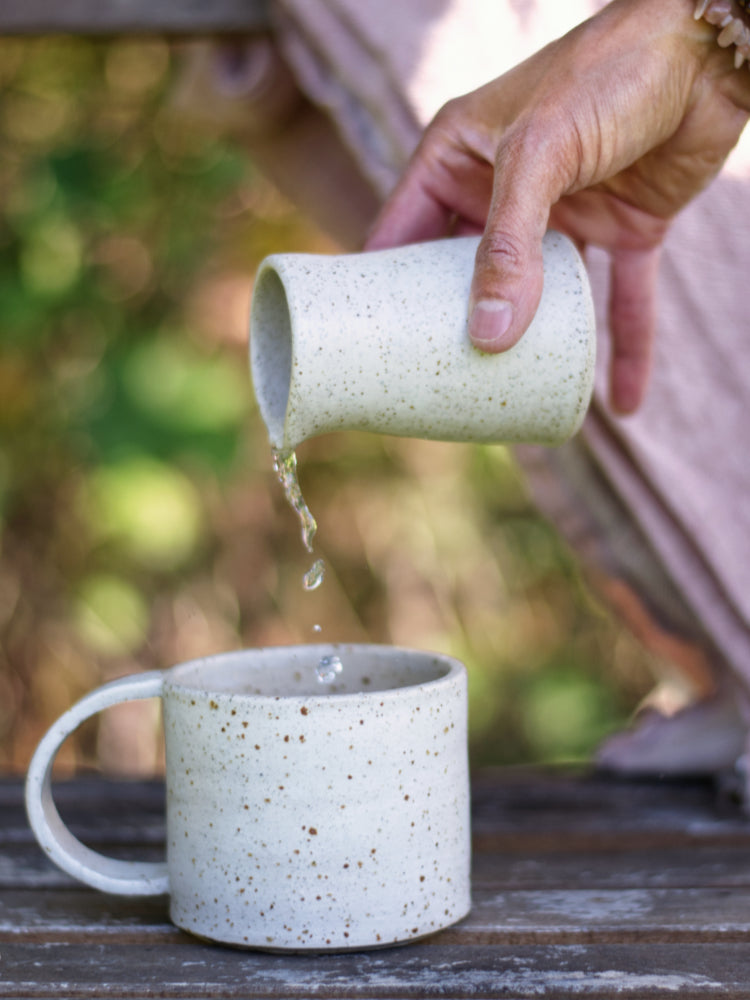Hand die een schenkkannetje vasthoudt en schenkt in een mok die staat op een stoel buiten.