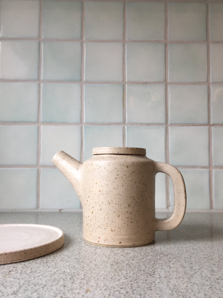 Handgemaakte theepot van gespikkelde klei naast een cakebord met een achtergrond van een tegelmuur met vierkante tegeltjes