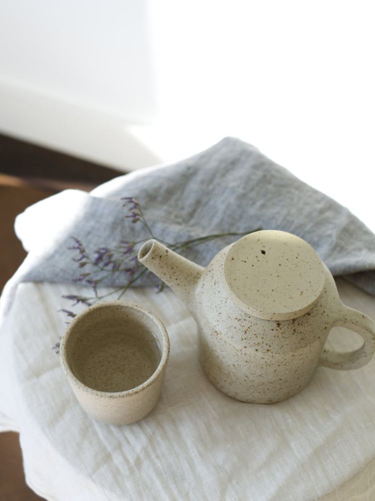 Rond laag tafeltje met daarop een handgemaakte theepot en kopje van gespikkelde klei naast een theedoek en een gedroogd takje met bloemetjes