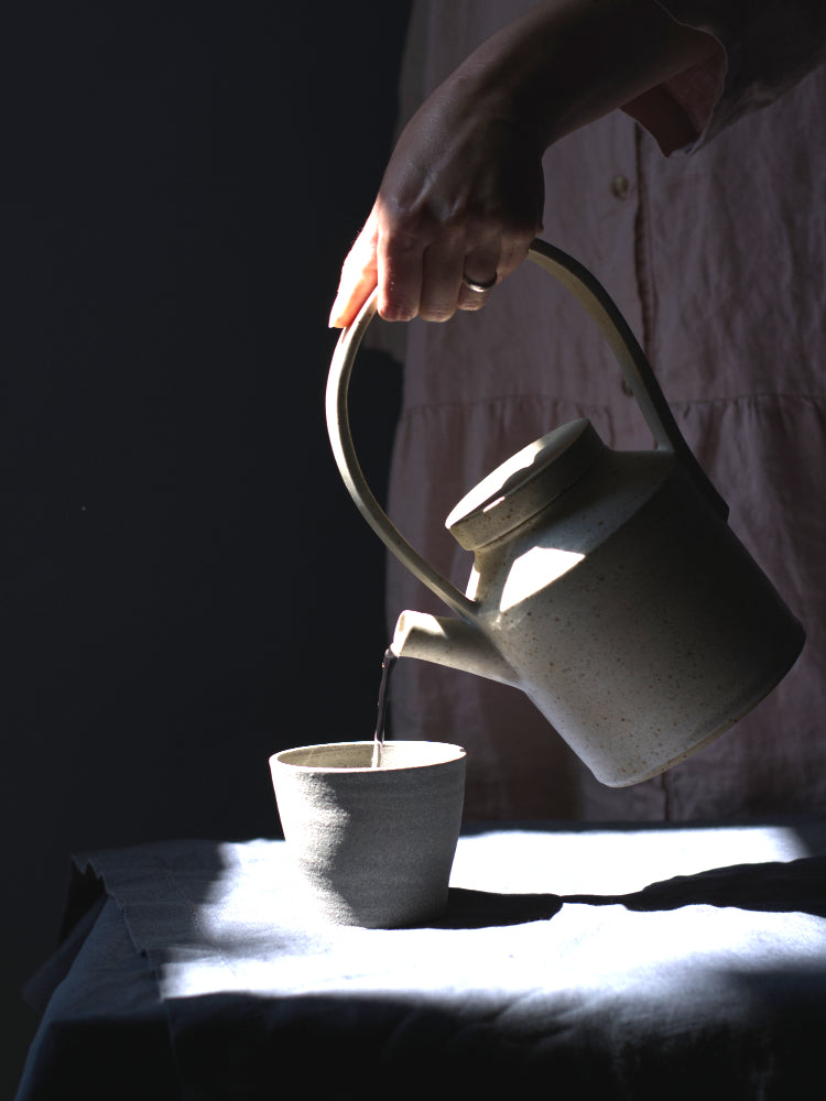 Handgemaakte theepot, vastgehouden door een hand, die schenkt in een theekopje, in het volle zonlicht met schadum eromheen