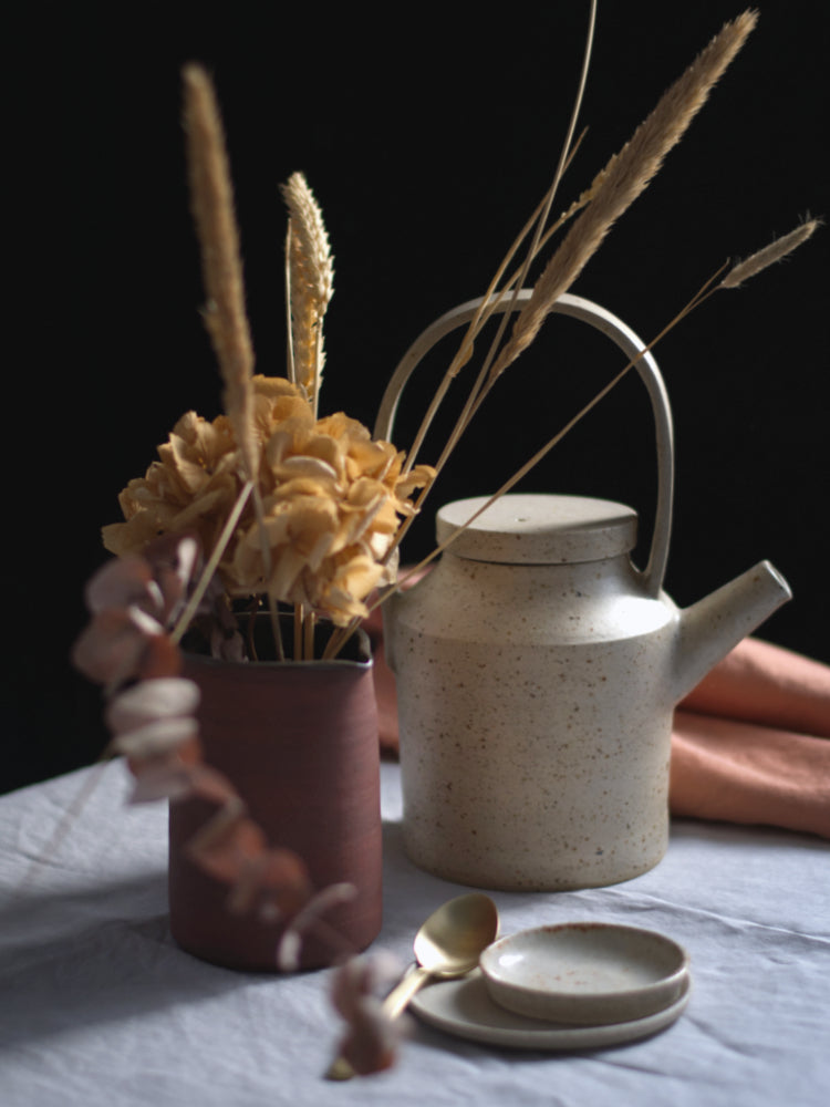 Handgemaakte theepot op een tafelkleed naast een schenkkannetje met droogbloemen.