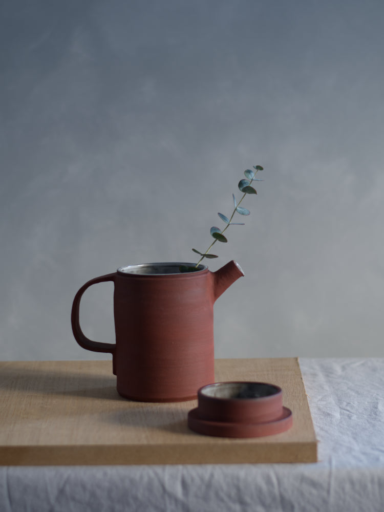 Handgemaakte rode theepot van steengoed op houten plank met een eucalyptustakje erin. Ernaast ligt de deksel van de theepot.