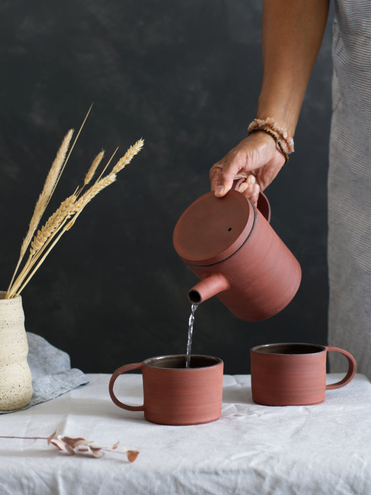 Hand die handgemaakte rode theepot vasthoudt en schenkt in mok van rood steengoed