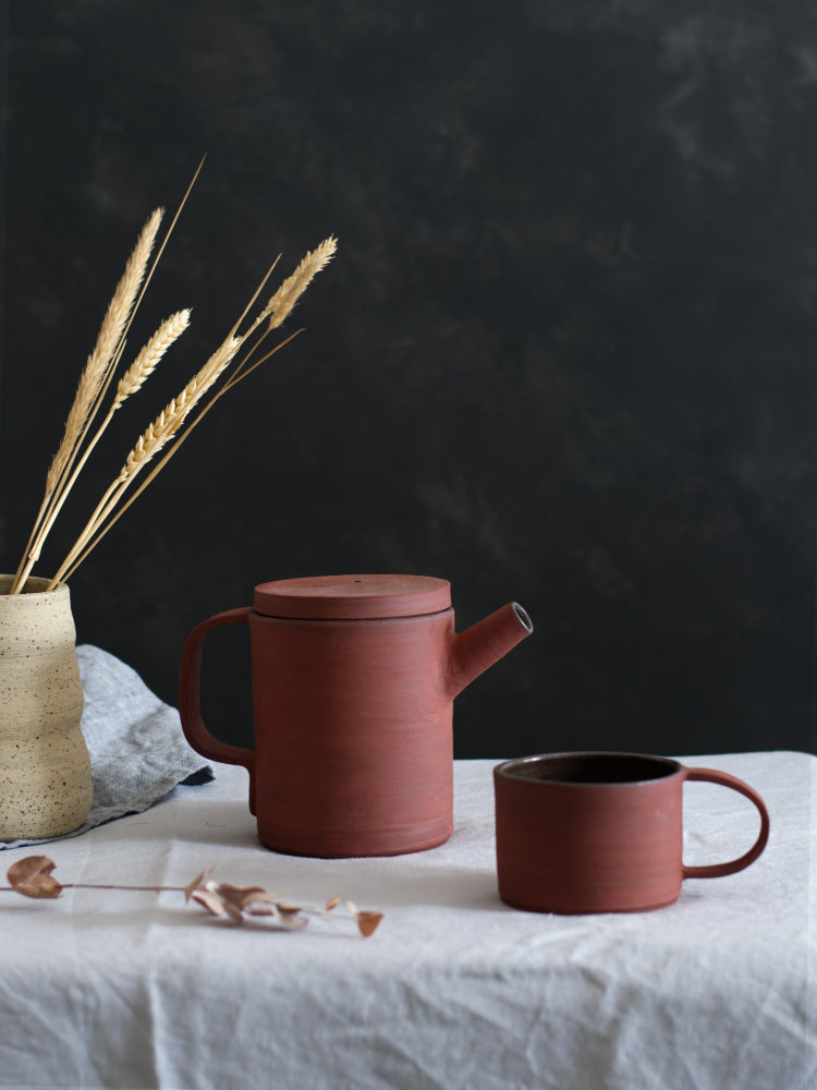 Handgemaakte rode theepot van steengoed op grijs tafelkleed naast rode mok en gespikkelde vaas met grashalmen