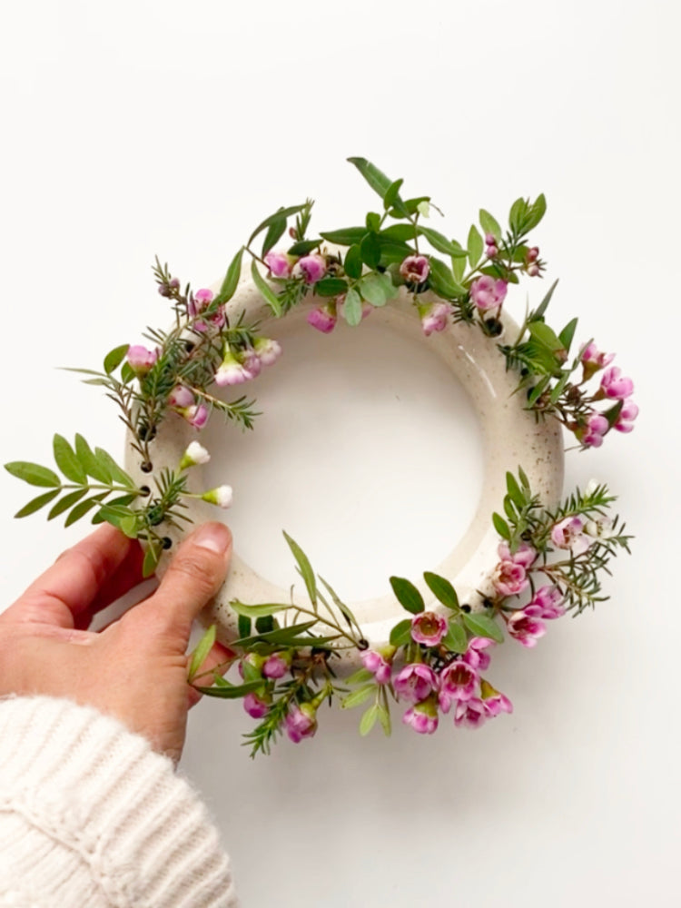 Ronde bloemenvaas met roze bloemen op een witte achtergrond en een handmet een witte wollige trui die hem vasthoudt.