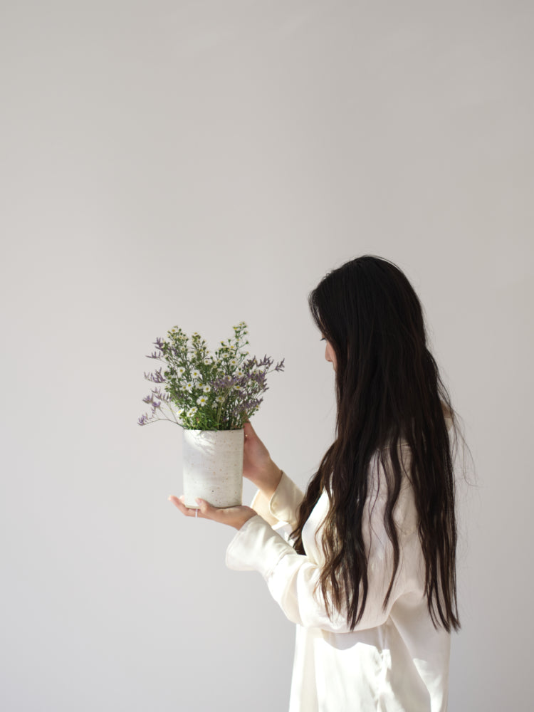 Bloemenvaas van keramiek, met bloemen, vastgehouden door een dame met lang donker haar en witte blouse.
