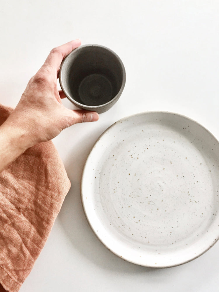 Whitewashed handgemaakt dinerbord op een witte achtergrond met een bruinroze theedoek ernaast en een hand die een kopje oppakt
