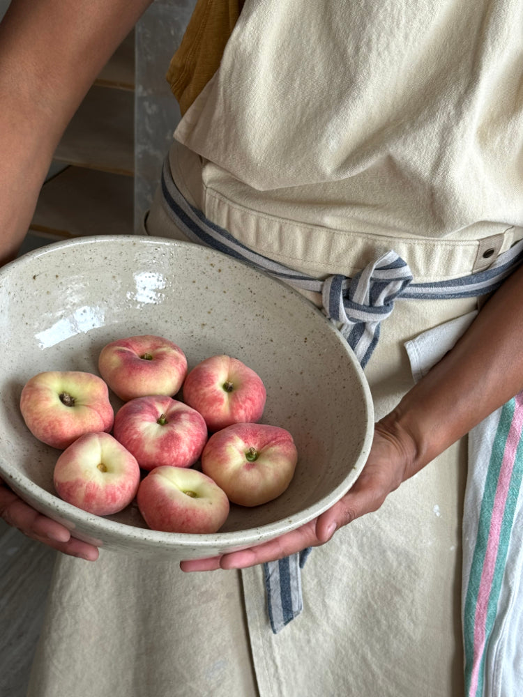 grote handgemaakte keramieken schaal met perziken, vastgehouden in handen met gestreept schort