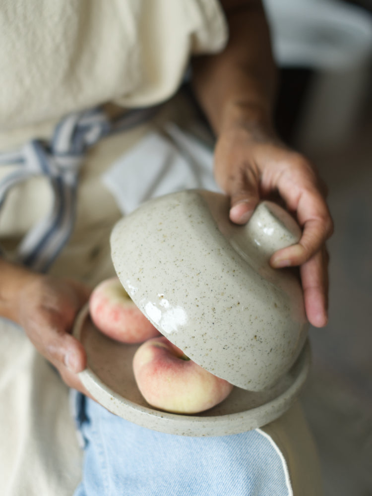 Handgemaakte keramieken stolp van gespikkelde klei, stolp wordt half opgetild door een hand, in de stolp zitten perziken. De stolp ligt op schoot bij iemand