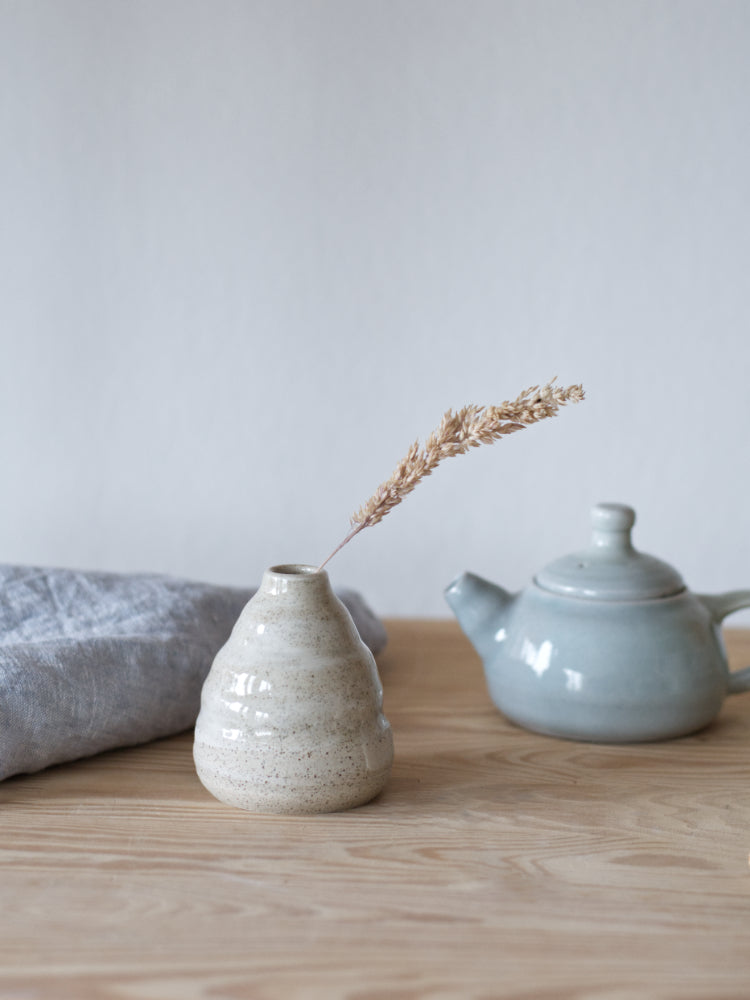 Handgemaakt klein vaasje, gemaakt van gespikkelde klei met licht golvende vorm met een grashalm naast een theedoek en een blauw theepotje