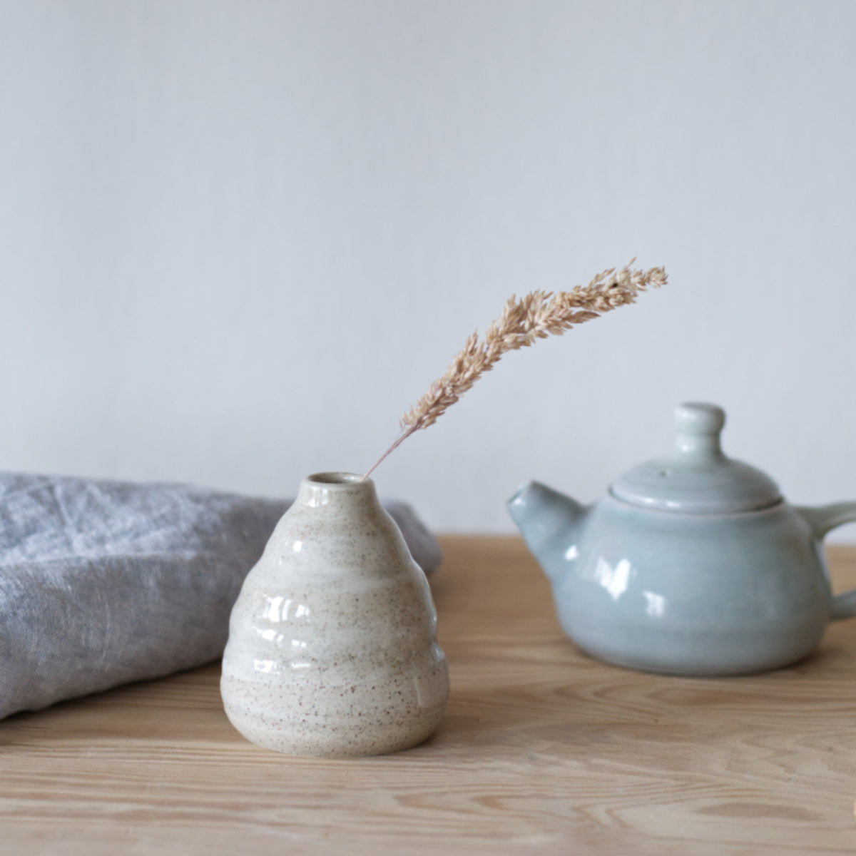 handgemaakt klein vaasje van gespikkeld keramiek met een grashalm en blauw theepotje en theedoek op de achtergrond