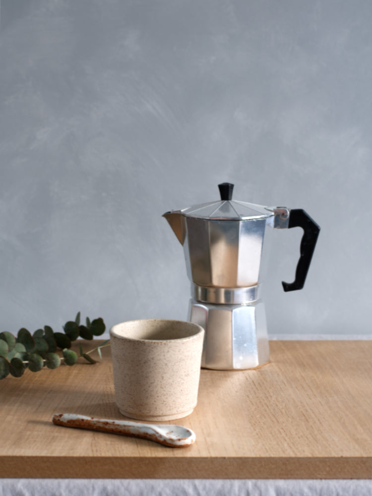 Handgemaakt kopje gespikkelde klei op een houten plank met lepel ervoor en percolator erachter