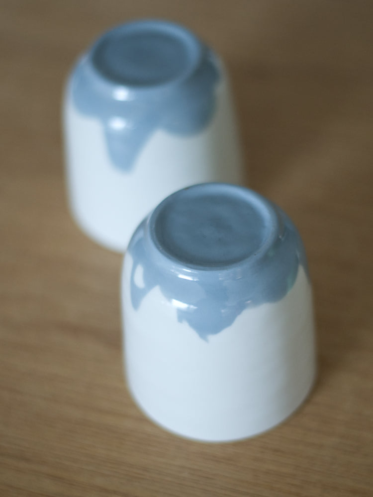 Twee handgemaakte Waterlijn kopjes medium van wit porselein met een blauwgrijze waterlijn aan de onderzijde gefotografeerd. Op een houten tafel als ondergrond