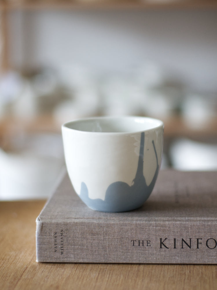 Handgemaakte Waterlijn kopje large van wit porselein met een grijsblauwe waterlijn, staand op een boek met linnen kaft, op een houten tafel