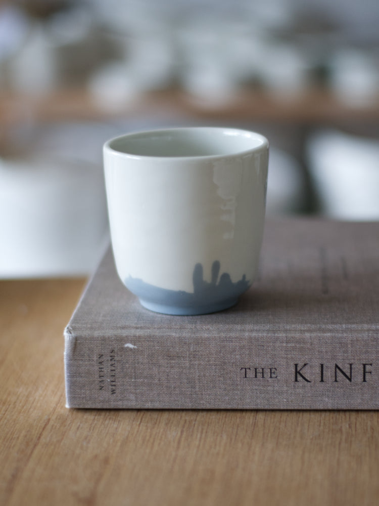 Handgemaakte Waterlijn kopje medium van wit porselein met een grijsblauwe waterlijn, staand op een boek met linnen kaft, op een houten tafel