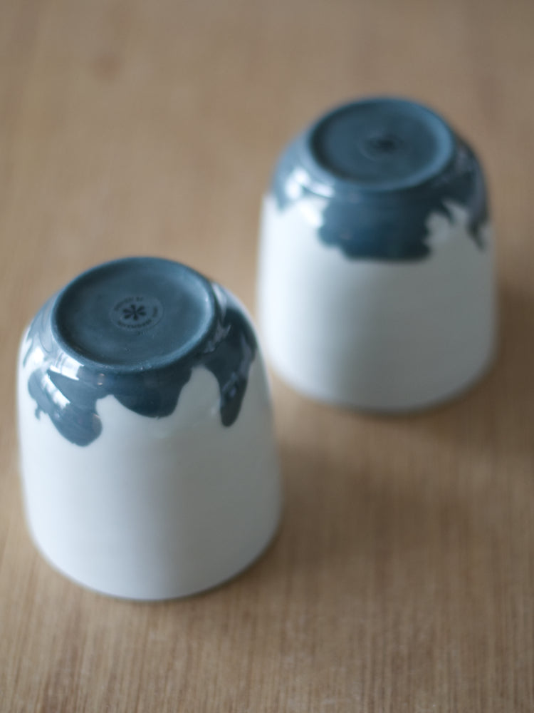 Twee handgemaakte Waterlijn kopjes medium van wit porselein met donkergrijze waterlijn aan de onderzijde gefotografeerd. Op een houten tafel als ondergrond