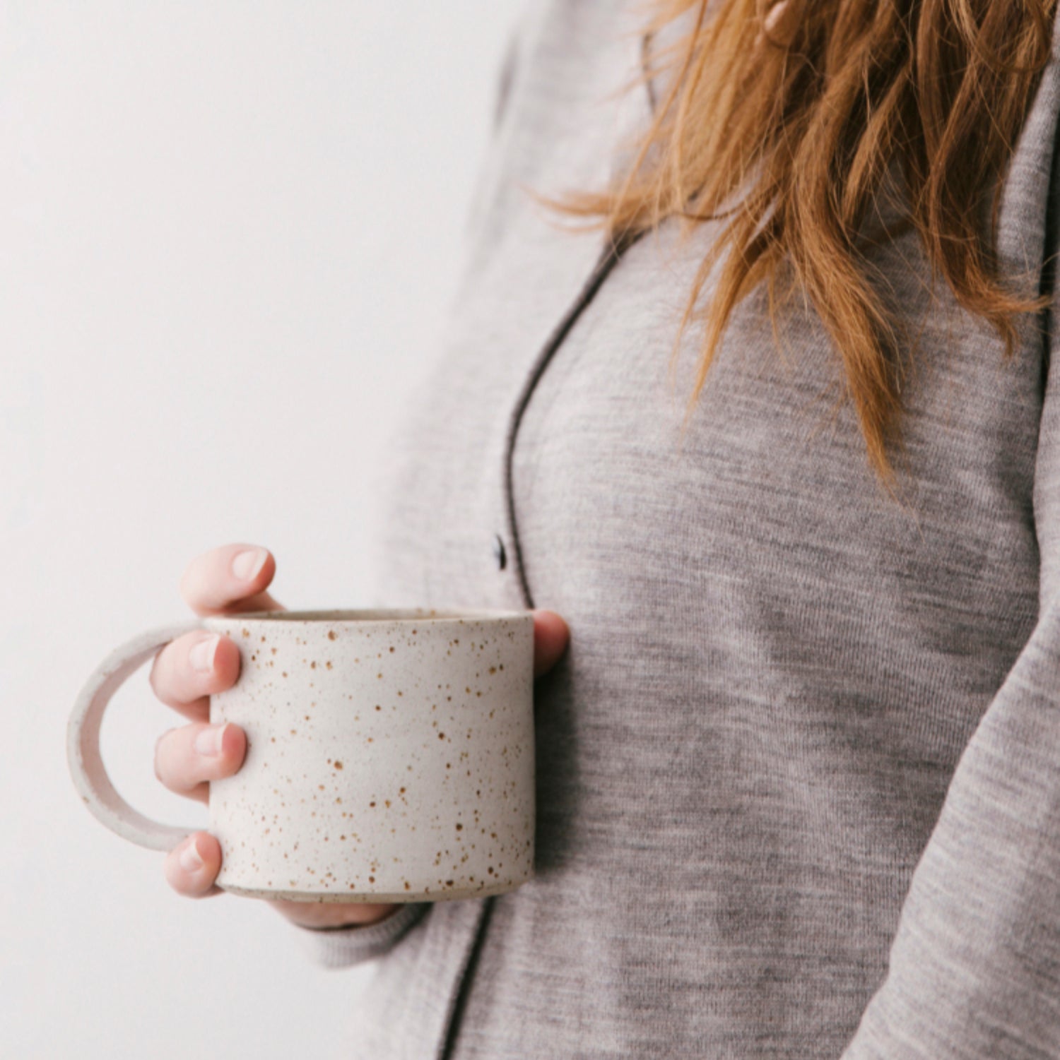 Handgemaakte whitewashed mok in een vrouwenhand. Je ziet alleen een pluk roodbruin haar en haar vest
