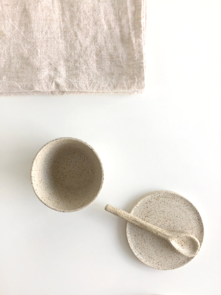 Handgemaakte lichtgespikkelde schotel en kopje van keramiek met lepel en een theedoek op een witte achtergrond