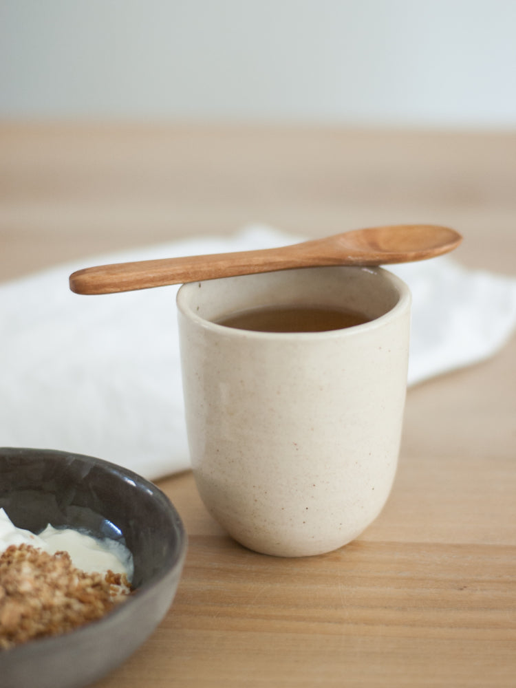 Handgemaakte theekop op een houten tafel met een houten lepel erop en een kommetje muesli ernaast