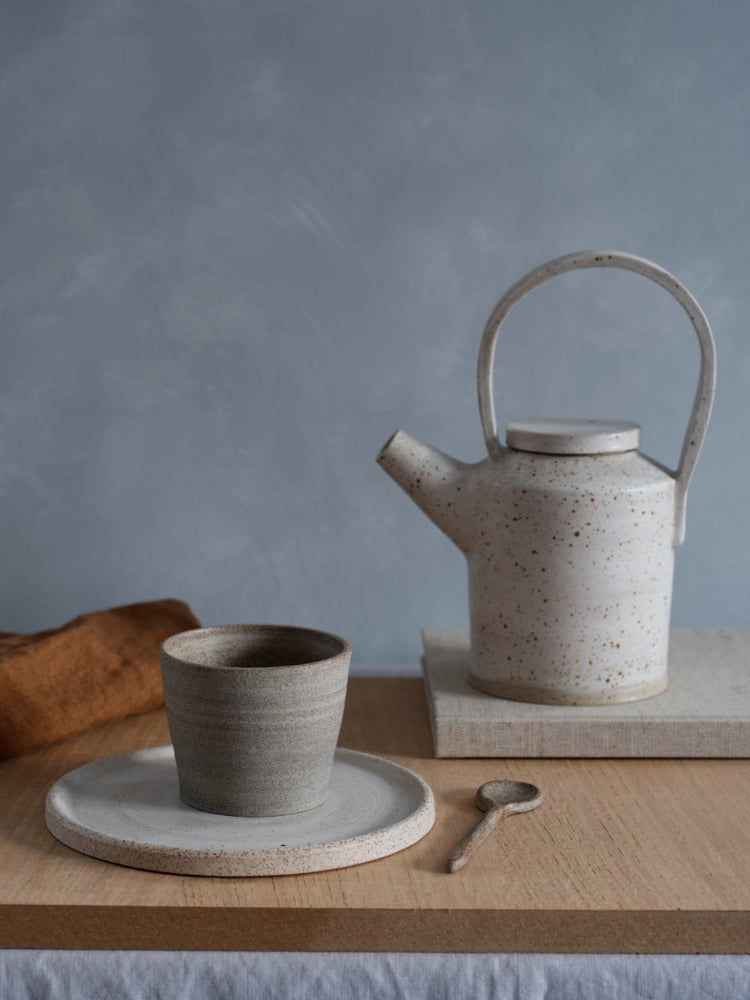 Handgemaakte keramieken theepot naast een cakebord met een kopje erop en een keramieken lepel ernaast op een houten plank