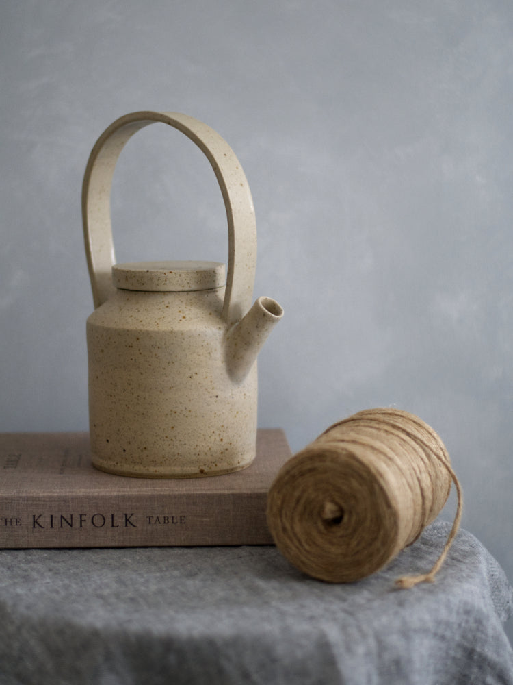 Handgemaakte theepot van gespikkelde klei op een boek op een krukje met theedoek en een rol touw