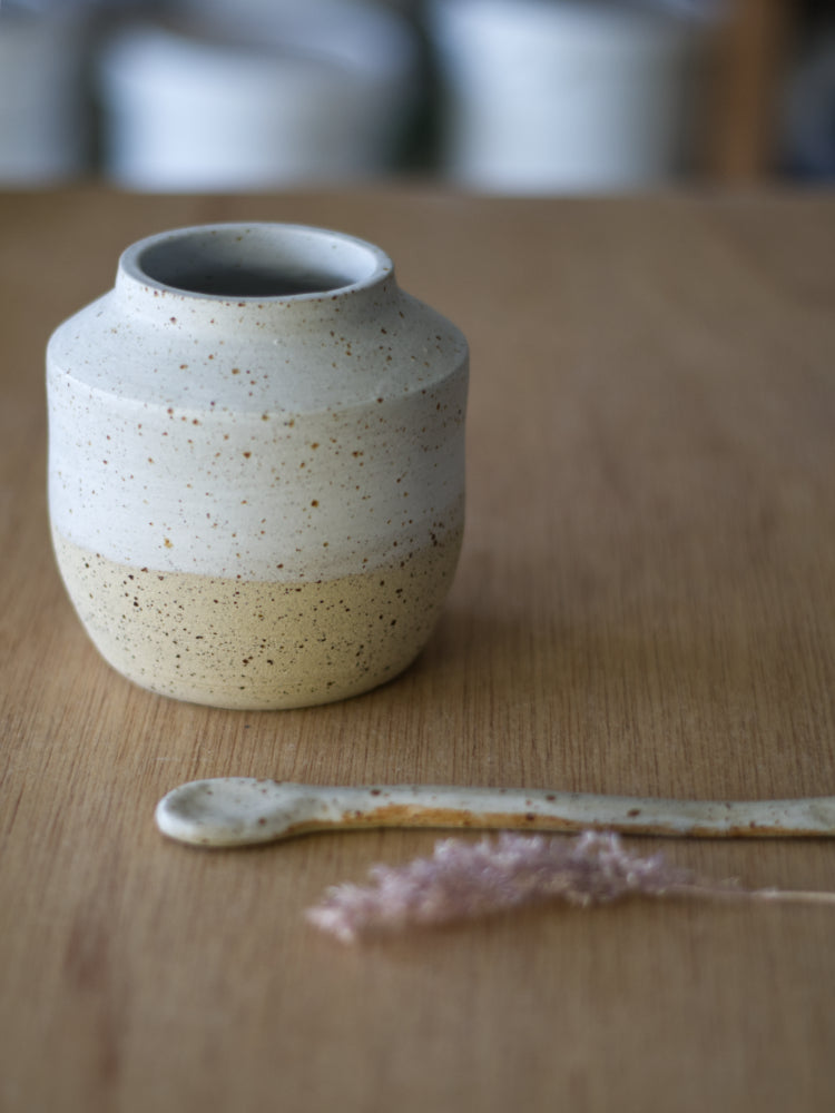 Handgemaakt vaasje van gespikkelde klei half wit mat geglazuurd naast een keramiek lepel en gedroogd graspluimpje