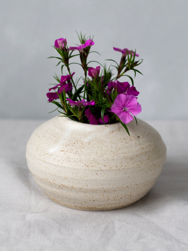 Handgemaakt vaasje gespikkelde klei met mat wit glazuur, met kleine roze bloemen erin op een lichtgrijs tafelkleed en grijze achtergrond