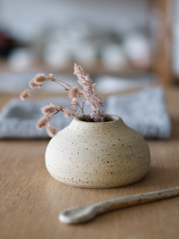 Handgemaakt vaasje gespikkelde klei met ongeglazuurde buitenkant met gedroogde bloemen erin op een houten tafel en keramiek lepel ernaast