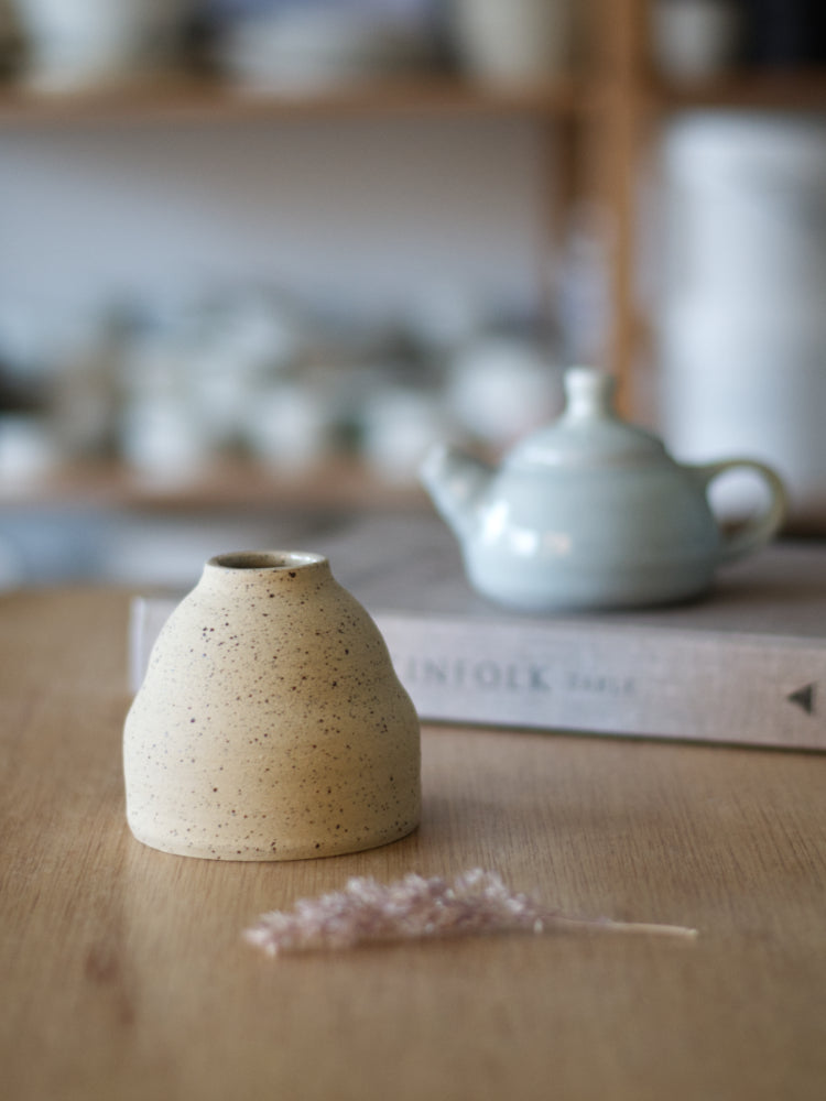 Handgemaakt klein vaasje van gespikkelde klei ongeglazuurde buitenkant en in de achtergrond een boek met een klein blauw theepotje erop