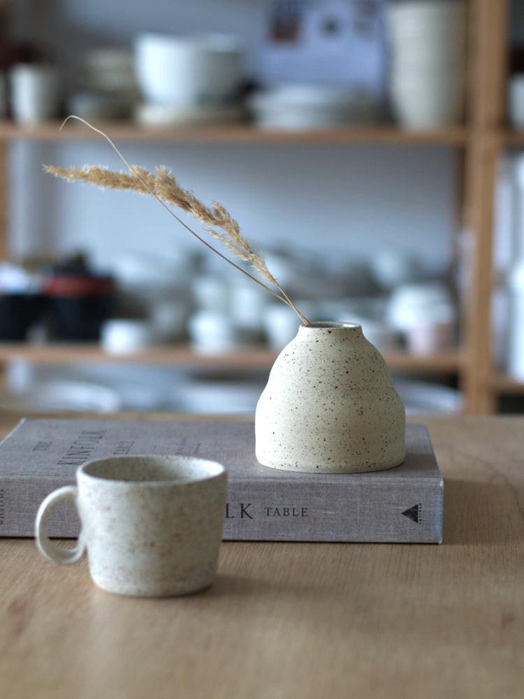 Handgemaakt vaasje van gespikkelde klei ongeglazuurde buitenkant op een kinfolk boek met een klein gespikkeld kopje ervoor