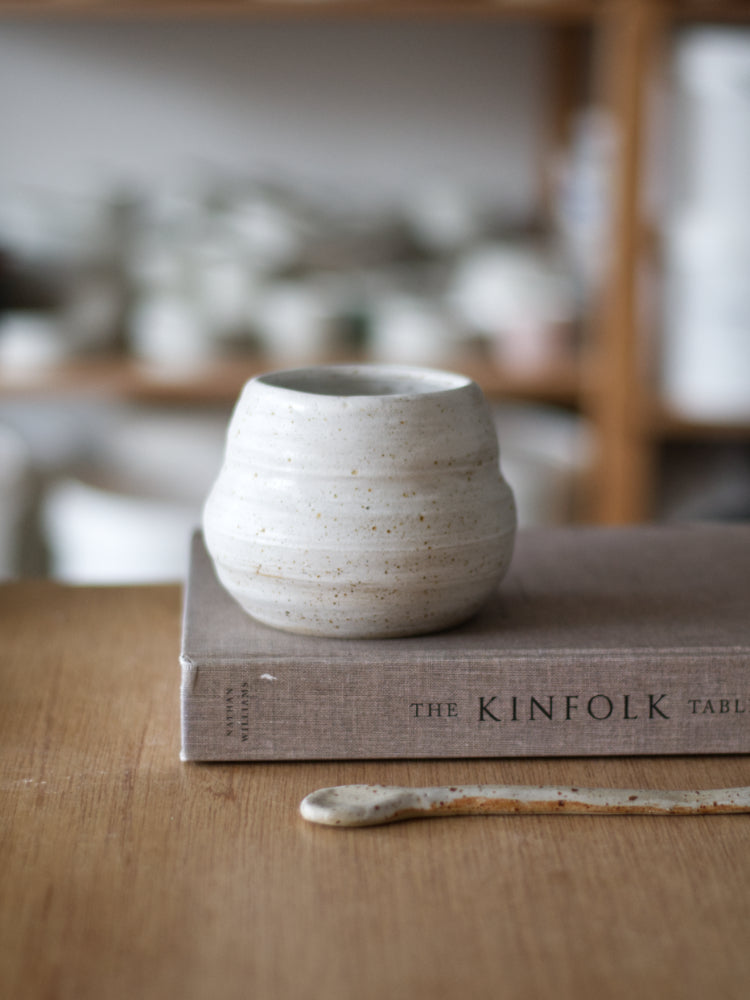 Handgemaakt klein vaasje van gespikkelde klei mat wit glazuur op een kinfolk boek met een keramiek lepel ervoor