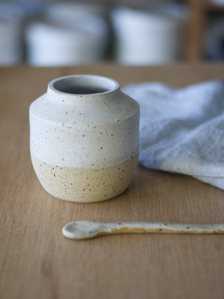 Handgemaakt vaasje van gespikkelde klei half wit mat geglazuurd naast een keramiek lepel, theedoek op de achtergrond