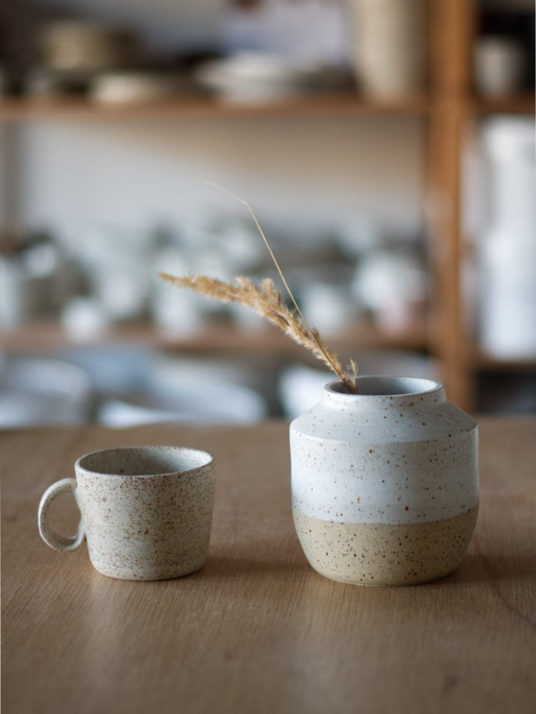 Handgemaakt vaasje van gespikkelde klei half wit mat geglazuurd naast een handgemaakt kopje
