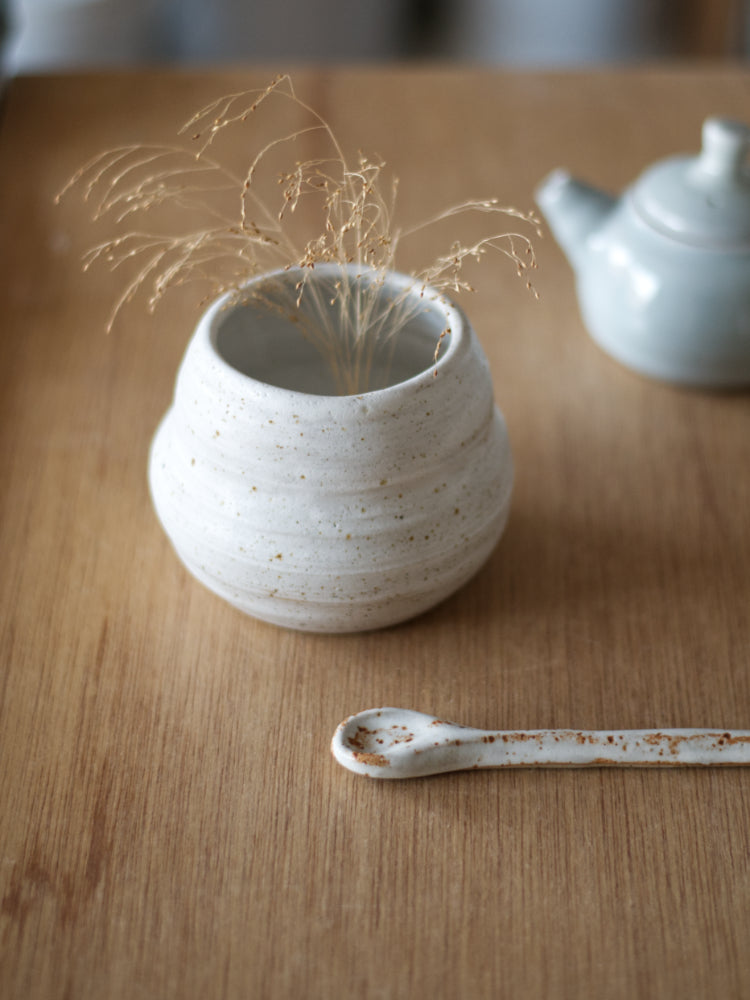 Handgemaakt klein vaasje van gespikkelde klei mat wit glazuur op een houten tafel met een keramiek lepel ervoor en een theepotje op de achtergrond