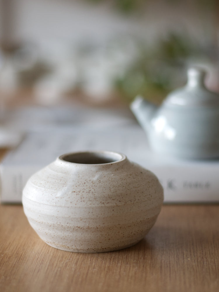 Handgemaakt vaasje met wit mat glazuur, met vaag erachter, op een boek, een klein blauw theepotje