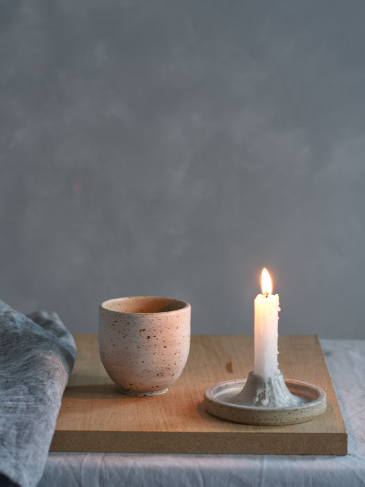 Handgemaakt kopje van gespikkelde klei, naast een brandende kaars in een handgemaakte kaarsenstandaard, grijze achtergrond, met een blauwe theedoek ernaast.