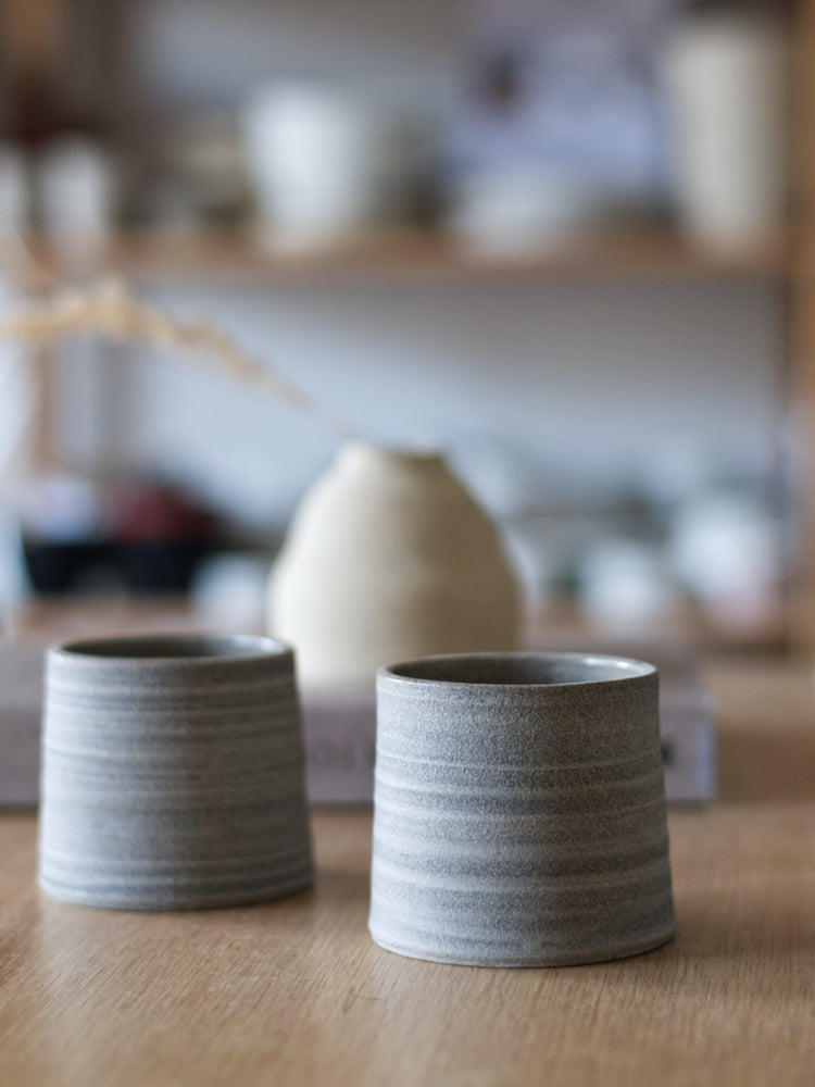 Donkere handgemaakte kopjes met ongeglazuurde buitenkant, met een vaag vaasje op de achtergrond