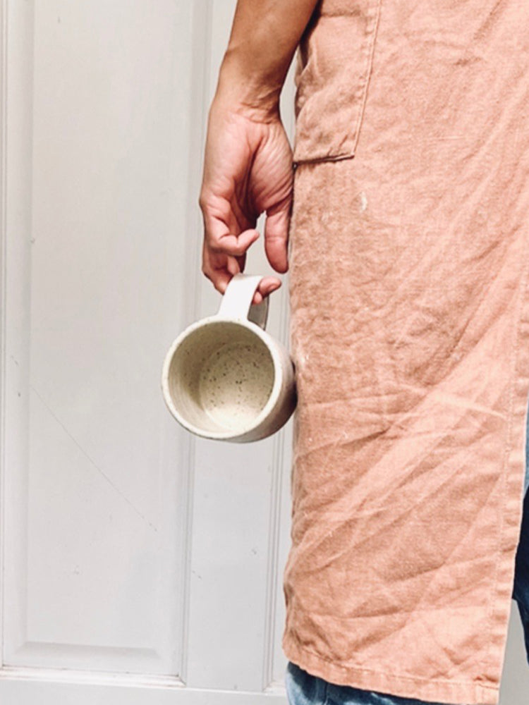 Iemand met een schort van achter gefotografeerd met in de hand een witgespikkelde handgemaakte mok die aan een paar vingers bungelt.