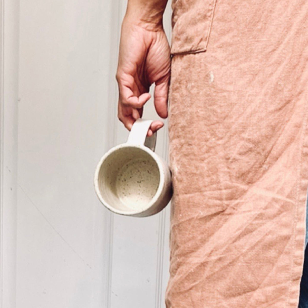 Close-up van een hand die een handgemaakte keramieken mok vasthoudt, met linnen schort