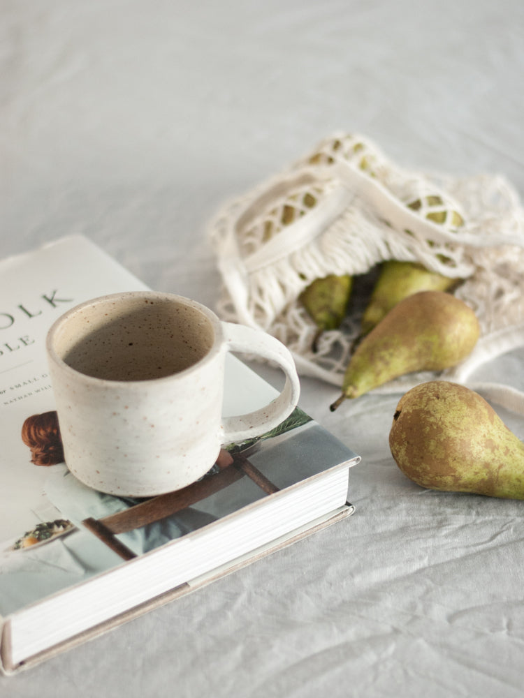 Handgemaakte witgespikkelde keramieken mok in slow living stijl op een boek met peren ernaast in een nettasje