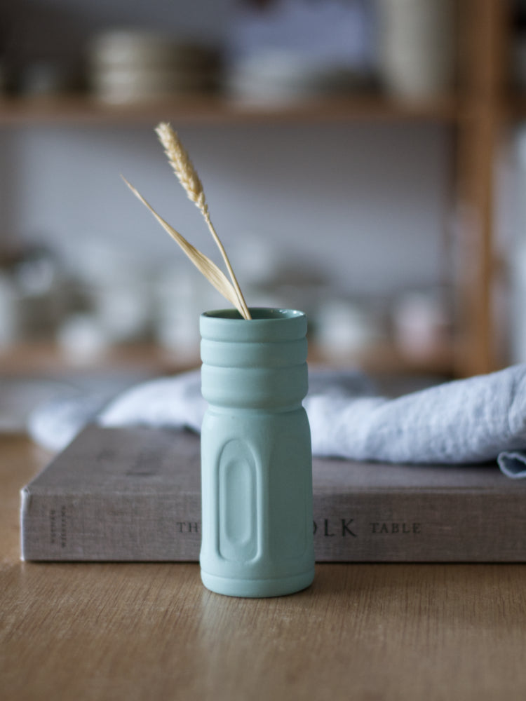 Handgemaakt groen porseleinen vaasje met boogpatroon op houten tafel naast een boek en theedoek