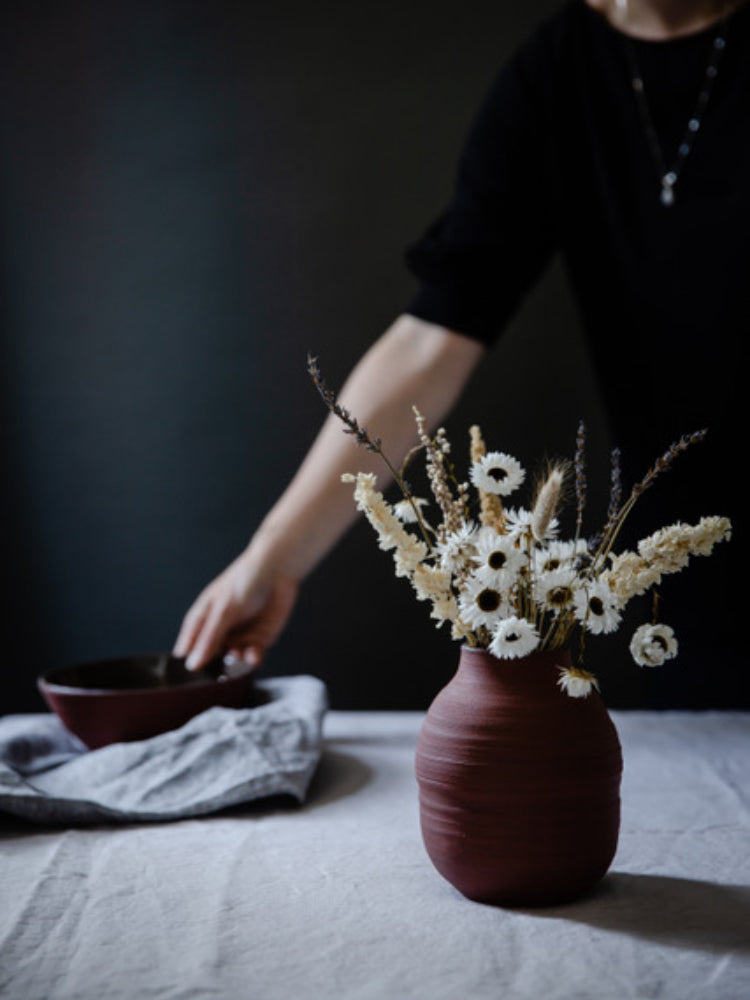 Rode bloemenvaas op een tafelkleed, met daarnaast een theedoek en iemand die een kom pakt. Donkere achtergrond.