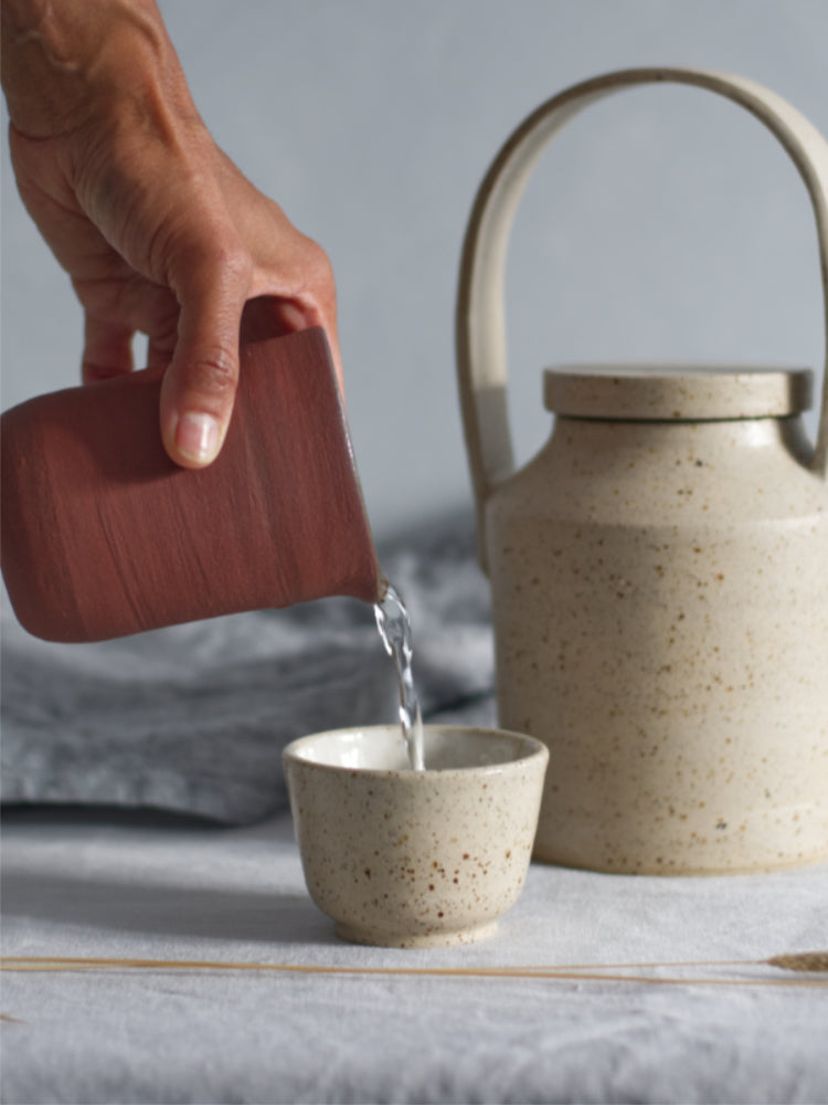 Rood schenkkannetje vastgehouden door een hand dat water schenkt in een kopje, op een tafelkleed met handgemaakte theepot ernaast.