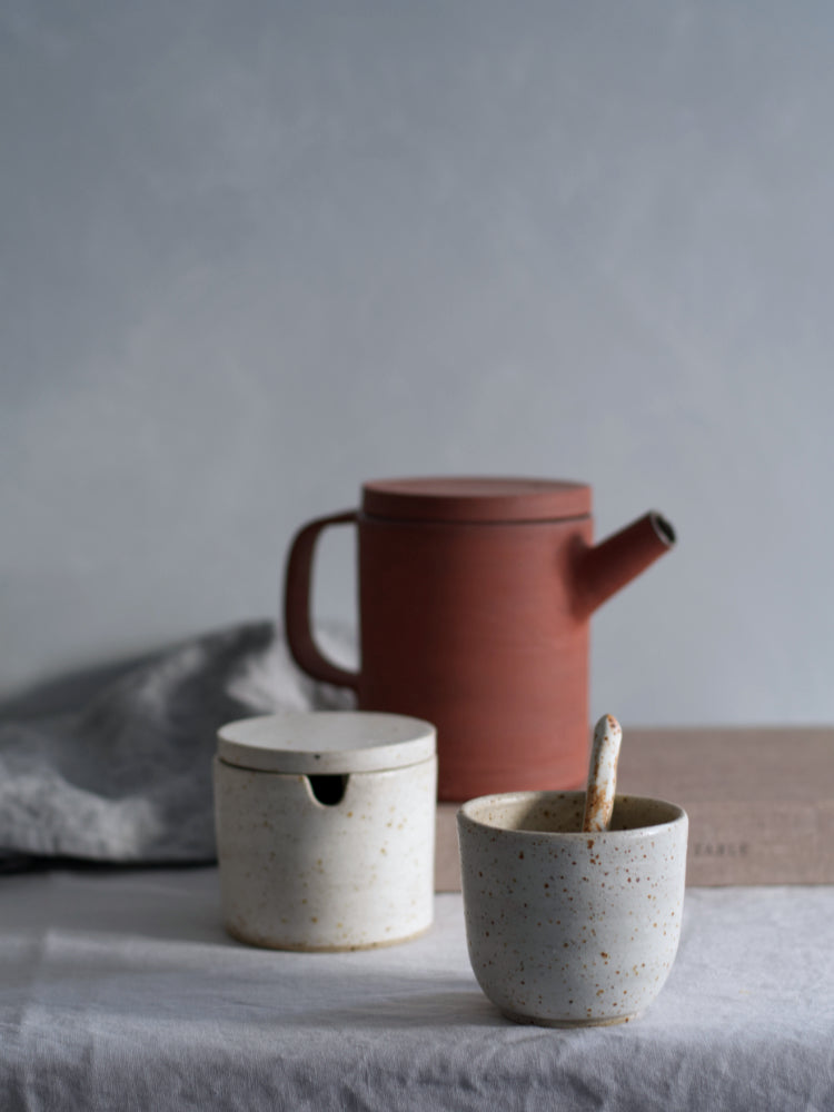 Handgemaakte rode theepot van steengoed op grijs tafelkleed naast suikerpotje en kopje van gespikkelde klei
